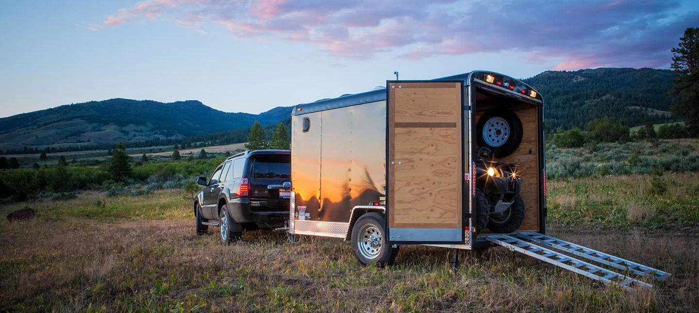 Black SUV towing a small enclosed cargo trailer with the rear ramp open and an ATV loaded inside, parked in a mountain meadow at sunset
