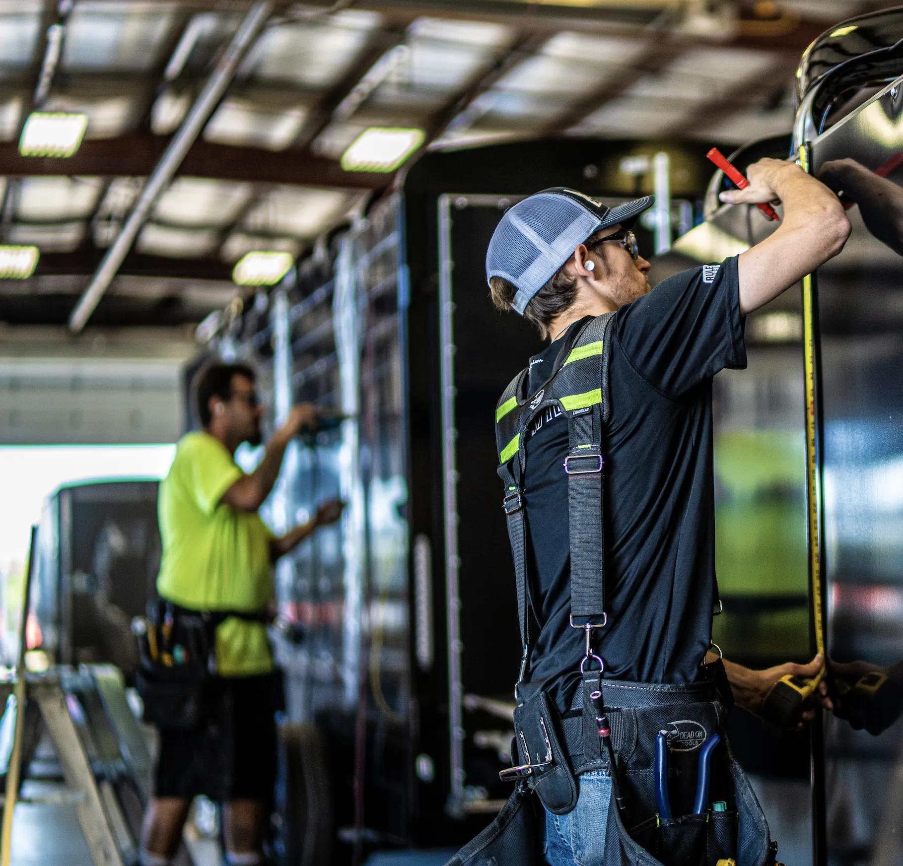 Two TrailersPlus technicians in safety harnesses and hi-vis gear measuring and inspecting a black trailer inside the service bay