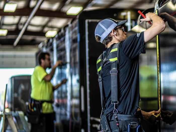 Two TrailersPlus technicians in safety harnesses and hi-vis gear measuring and inspecting a black trailer inside the service bay