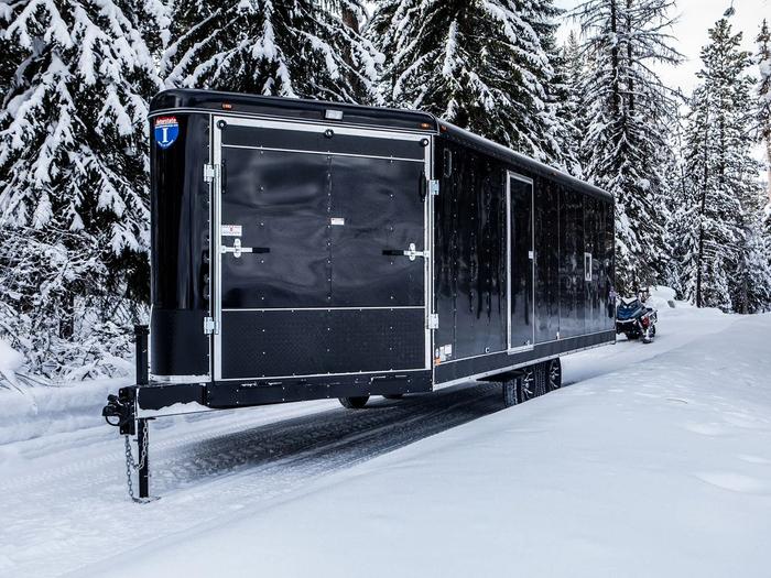Black Interstate enclosed snowmobile trailer parked on a snow-covered forest road with a rider on a snowmobile beside it
