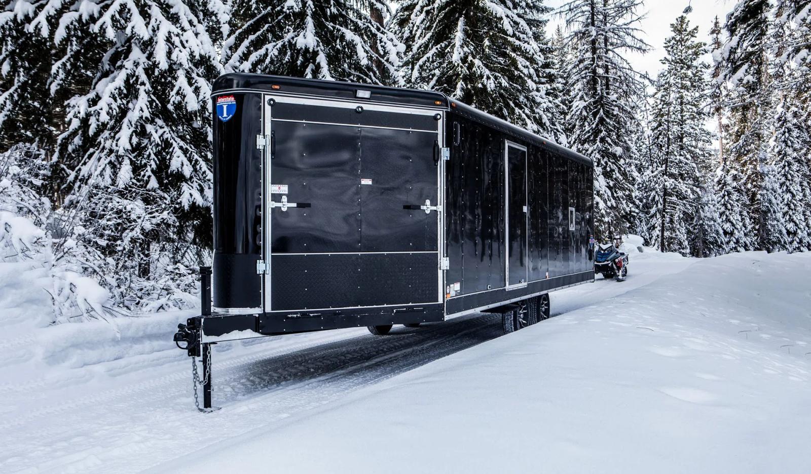 Black Interstate enclosed snowmobile trailer parked on a snow-covered forest road with a rider on a snowmobile beside it