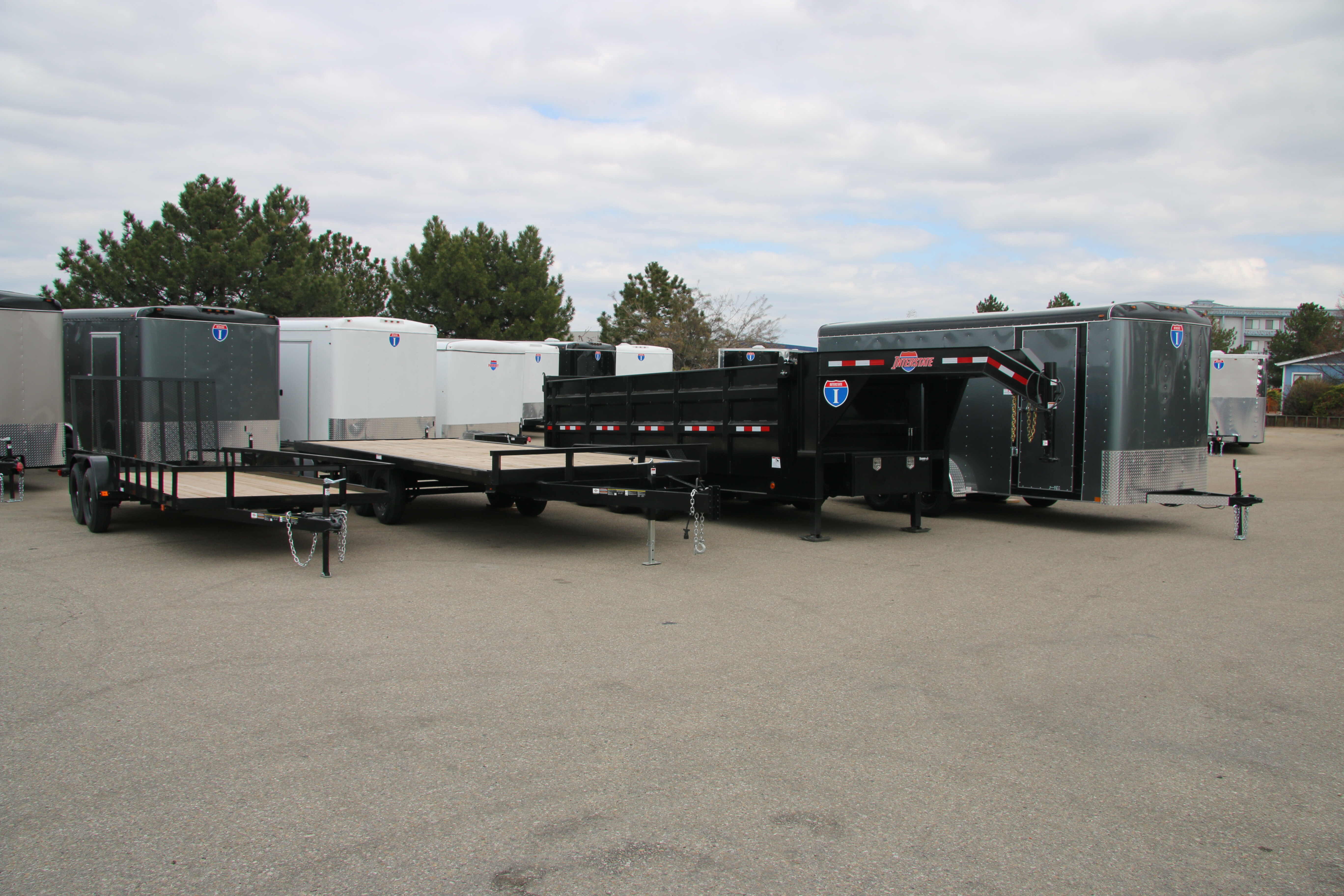 Lineup of a black utility, flatbed deckover, gooseneck dump, and charcoal enclosed cargo trailer with white cargo trailers behind on a TrailersPlus lot