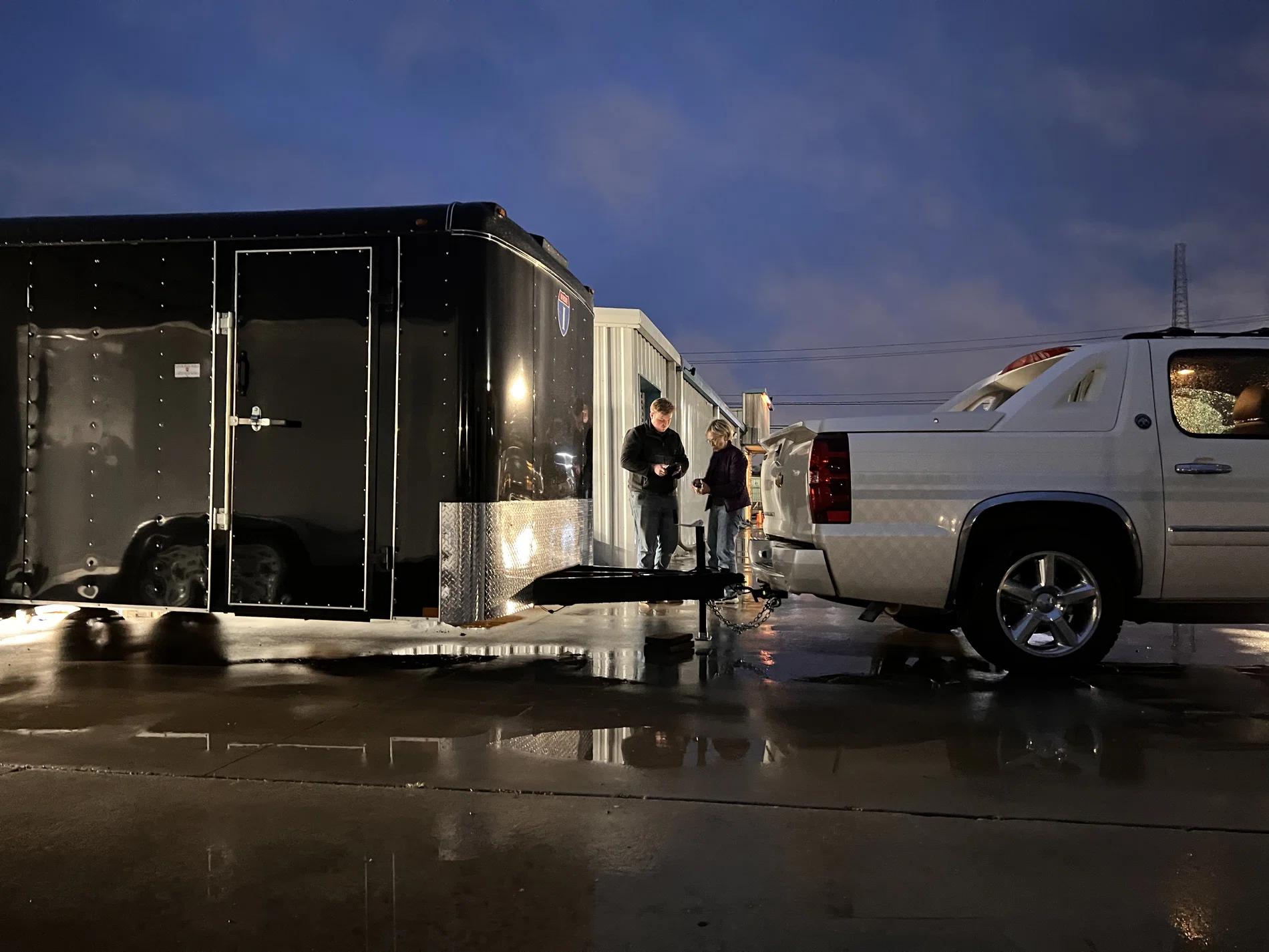 Black enclosed cargo trailer with side door open and interior lights on, hitched behind a white SUV on a wet lot at dusk