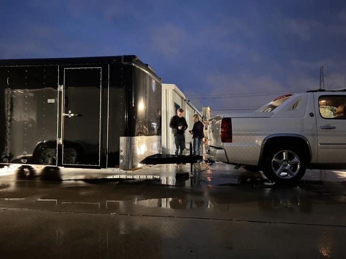 Black enclosed cargo trailer with side door open and interior lights on, hitched behind a white SUV on a wet lot at dusk