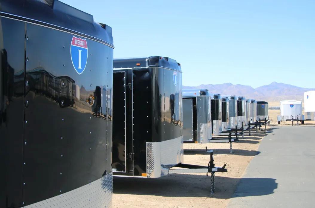 Receding row of alternating black and silver enclosed cargo trailers parked along a paved drive at a desert dealer lot