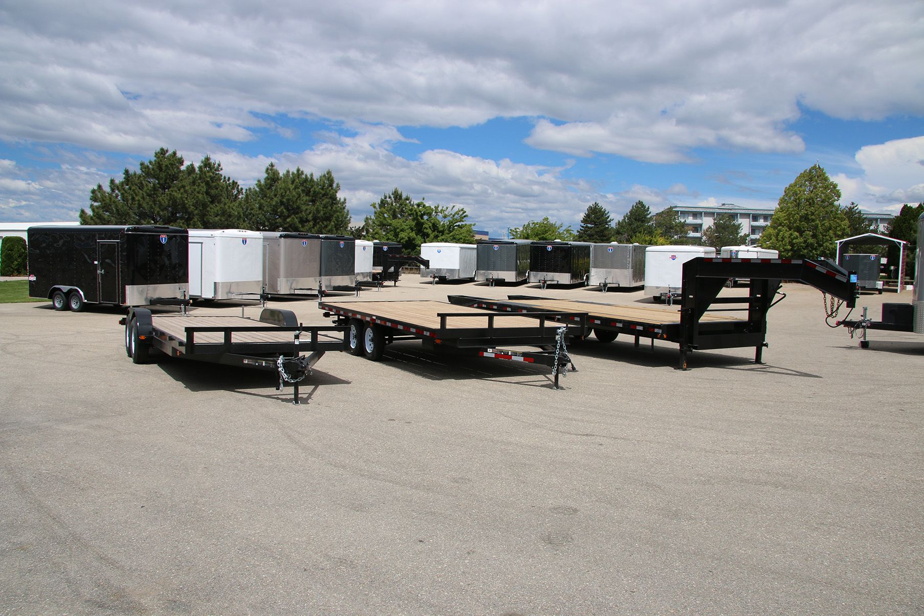 TrailersPlus dealer lot with two flatbed deckover gooseneck trailers in front of long rows of black and white enclosed cargo trailers
