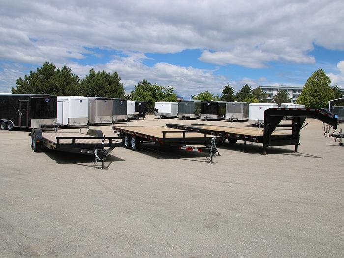 TrailersPlus dealer lot with two flatbed deckover gooseneck trailers in front of long rows of black and white enclosed cargo trailers