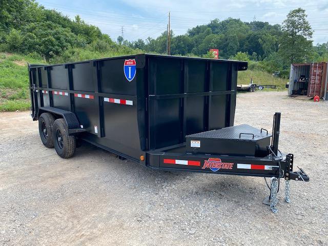 Black tandem-axle utility trailer with wood deck and rear mesh ramp gate parked on gravel in front of a blue house and red barn