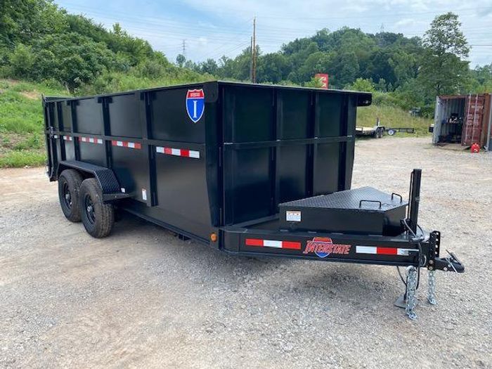 Black tandem-axle utility trailer with wood deck and rear mesh ramp gate parked on gravel in front of a blue house and red barn