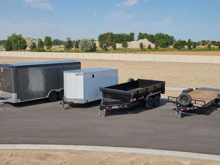 Lineup of charcoal and white enclosed cargo, black dump, and flatbed utility trailers parked on asphalt at a TrailersPlus lot