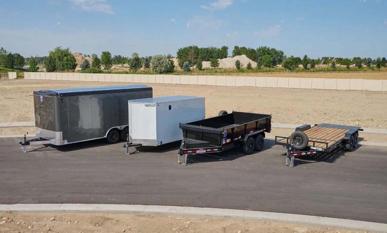 Lineup of charcoal and white enclosed cargo, black dump, and flatbed utility trailers parked on asphalt at a TrailersPlus lot