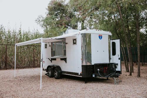 Group of attendees seated at picnic tables in front of a black TrailersPlus mobile event trailer at an outdoor tailgate gathering