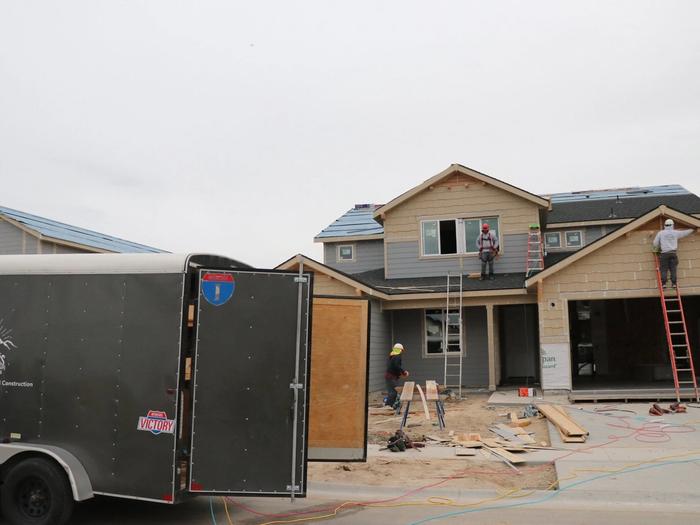 Gray enclosed cargo trailer with rear doors open at a residential construction site where workers in hard hats frame a two-story house