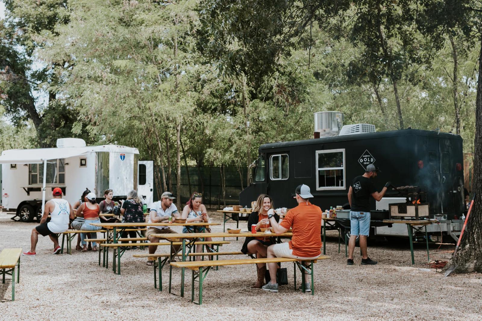 Group of attendees seated at picnic tables in front of a black TrailersPlus mobile event trailer at an outdoor tailgate gathering