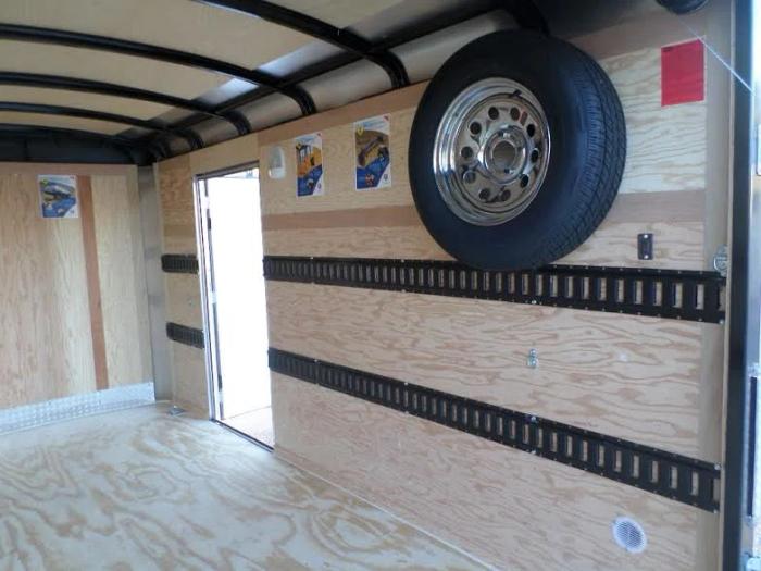 Interior of an enclosed cargo trailer with plywood walls, mounted E-track tie-down rails, and a spare tire mounted on the side wall