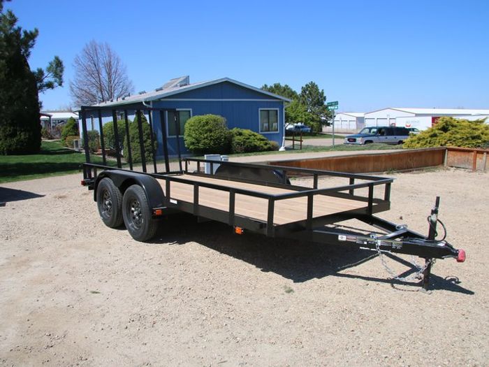 Rows of black single-axle utility trailers with wood decks lined up on a desert TrailersPlus dealer lot with a US flag and mountains in the distance