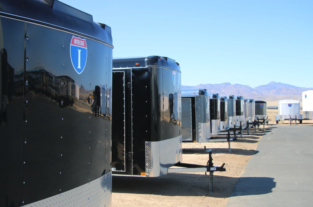 Receding row of alternating black and silver enclosed cargo trailers parked along a paved drive at a desert dealer lot