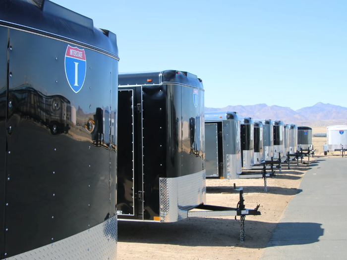 Receding row of alternating black and silver enclosed cargo trailers parked along a paved drive at a desert dealer lot