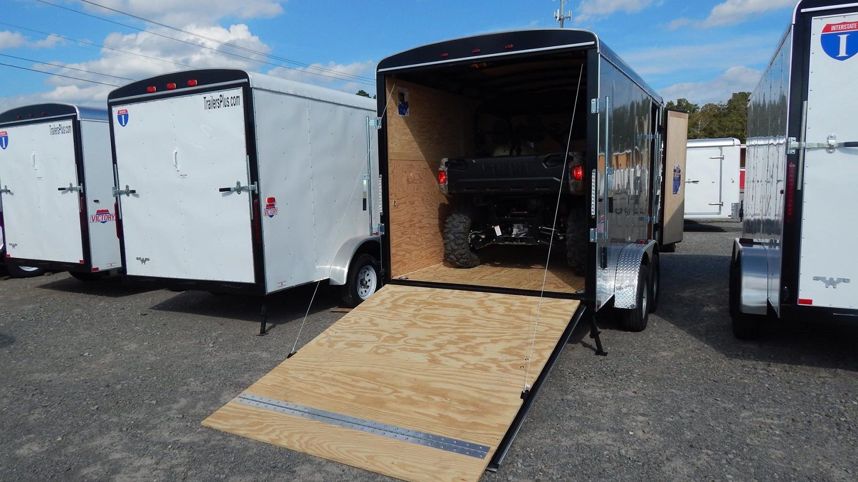 Lineup of a black utility trailer, flatbed deckover, gooseneck dump, and enclosed cargo trailers parked together on a TrailersPlus dealer lot under cloudy skies