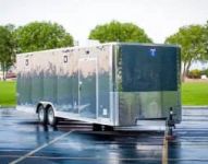 Lineup of a black utility trailer, flatbed deckover, gooseneck dump, and enclosed cargo trailers parked together on a TrailersPlus dealer lot under cloudy skies