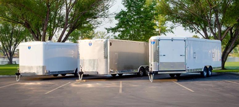 Three white enclosed cargo trailers parked at angled spaces in a tree-lined paved lot