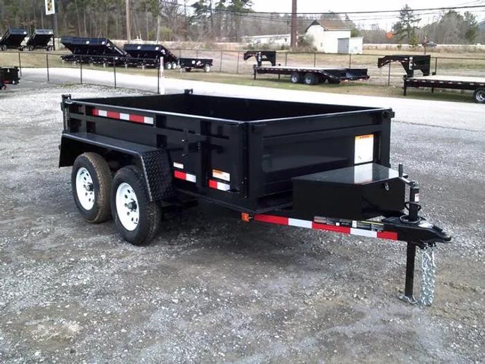 Black tandem-axle dump trailer parked on gravel at a TrailersPlus lot with rows of dump trailers and a fence behind