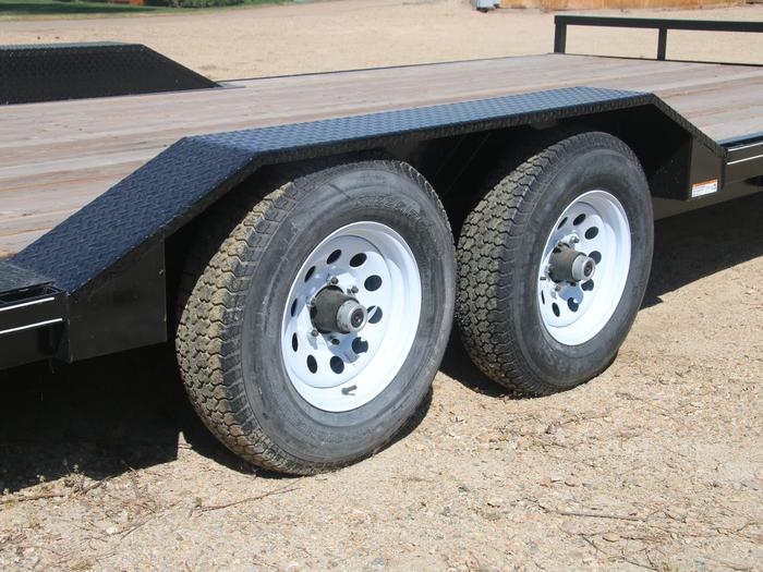 Close-up of a black equipment trailer's tandem axles with silver wheels and the edge of its wood plank deck