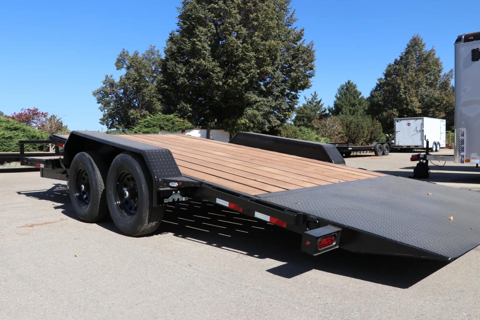 Black tandem-axle flatbed equipment trailer with rear loading ramp deployed exposing the wood deck, parked at a sunny dealer lot