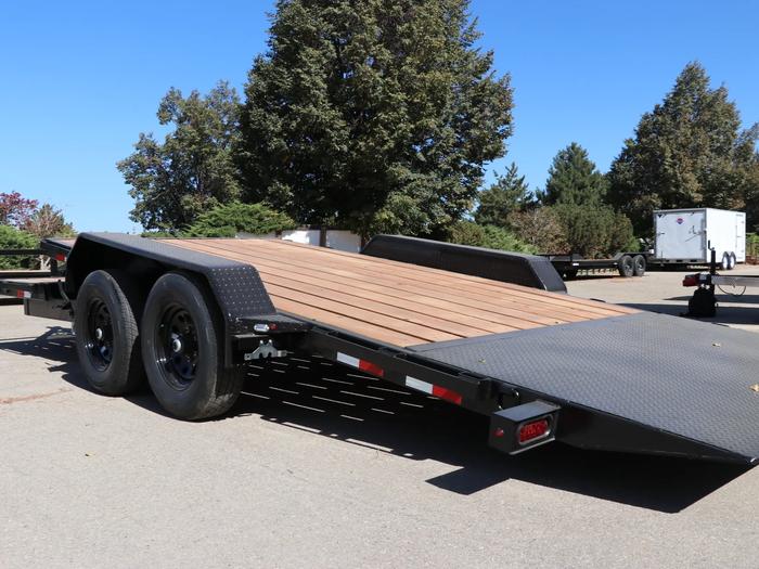 Black tandem-axle flatbed equipment trailer with rear loading ramp deployed exposing the wood deck, parked at a sunny dealer lot