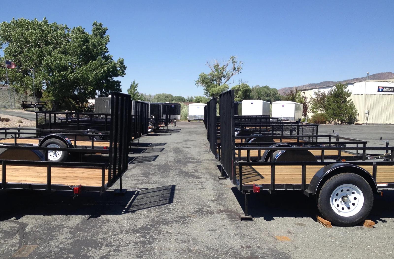 Two flatbed deckover gooseneck trailers parked in front of rows of enclosed cargo trailers at a TrailersPlus dealer lot