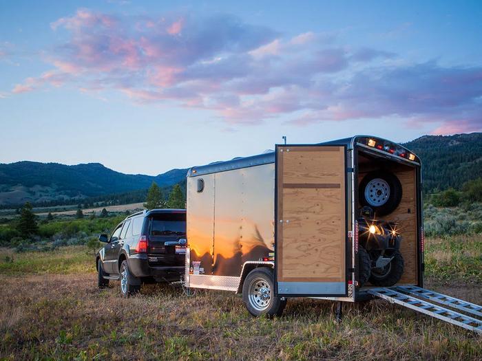 Black SUV towing a small enclosed cargo trailer with the rear ramp open and an ATV loaded inside in a mountain meadow at sunset