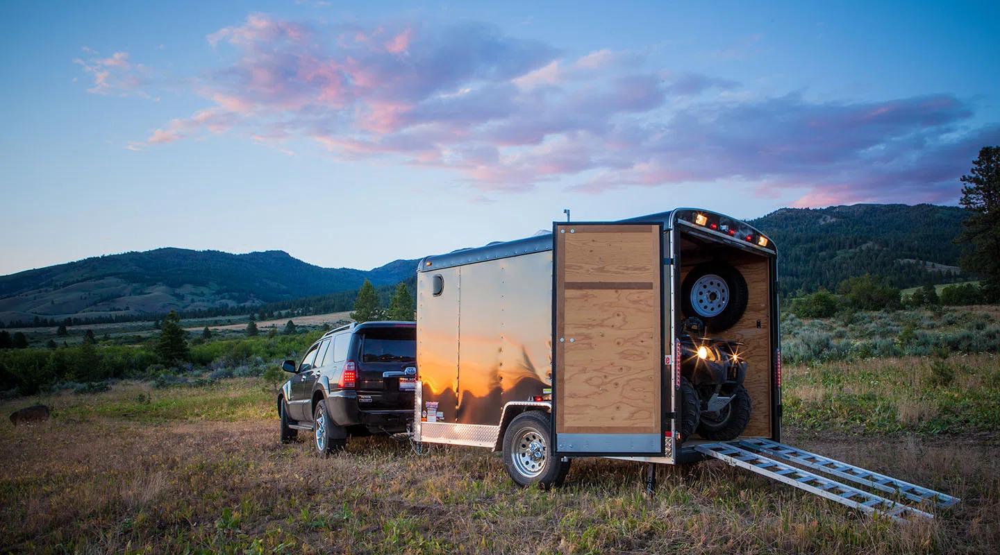 Black SUV towing a small enclosed cargo trailer with the rear ramp open and an ATV loaded inside in a mountain meadow at sunset