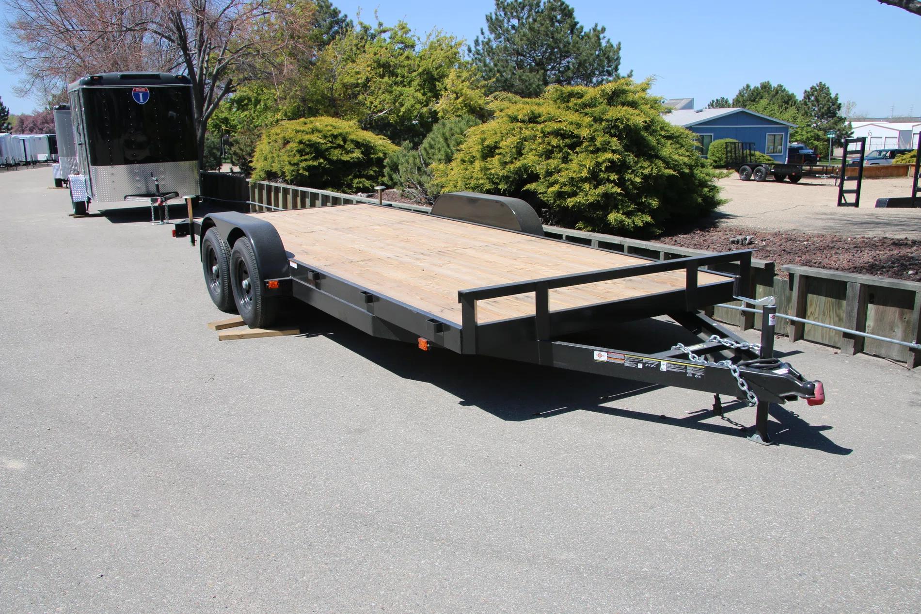 Black tandem-axle flatbed deckover equipment trailer with wood deck parked at a paved lot beside an enclosed cargo trailer