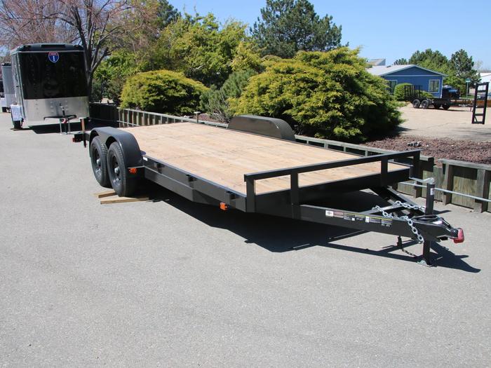 Black tandem-axle flatbed deckover equipment trailer with wood deck parked at a paved lot beside an enclosed cargo trailer