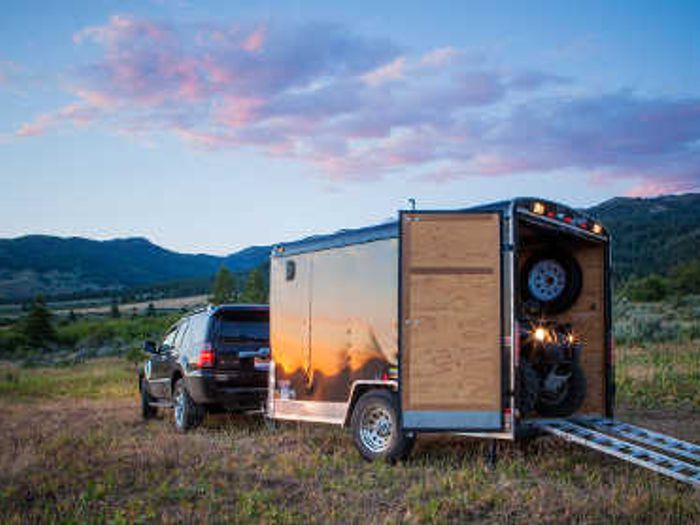 Black SUV towing a small enclosed cargo trailer with the rear ramp open and an ATV loaded inside, parked in a mountain meadow at sunset