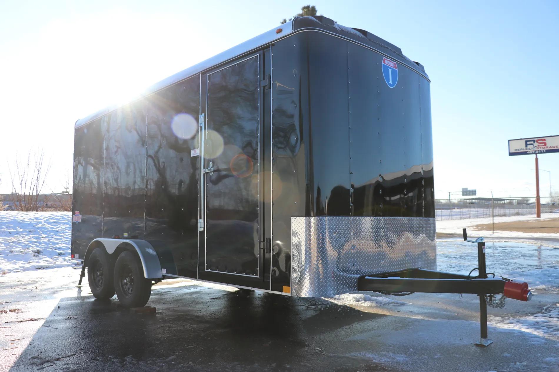 Black Interstate enclosed cargo trailer with diamond-plate guard parked on a sunlit icy asphalt lot with patches of snow