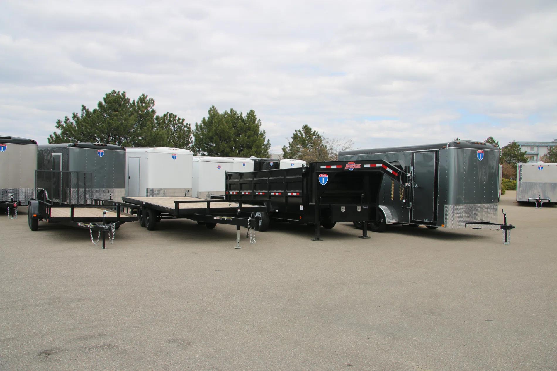 Lineup of a black utility trailer, flatbed deckover, gooseneck dump, and enclosed cargo trailers parked together on a TrailersPlus dealer lot under cloudy skies