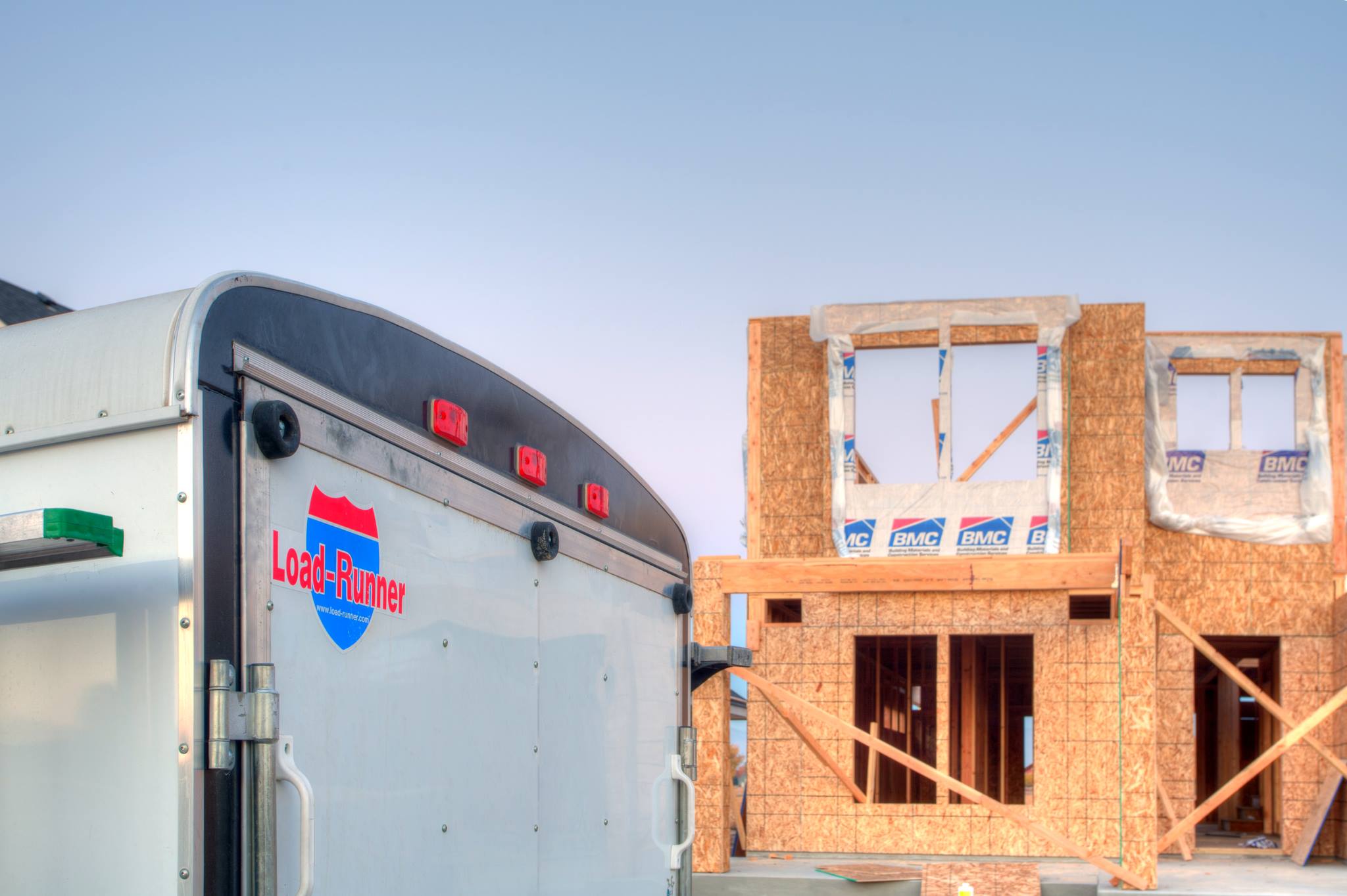 Rear of a white enclosed cargo trailer with a Load-Runner shield decal, parked beside a wood-framed house under construction