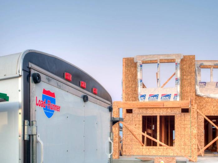 Rear of a white enclosed cargo trailer with a Load-Runner shield decal, parked beside a wood-framed house under construction