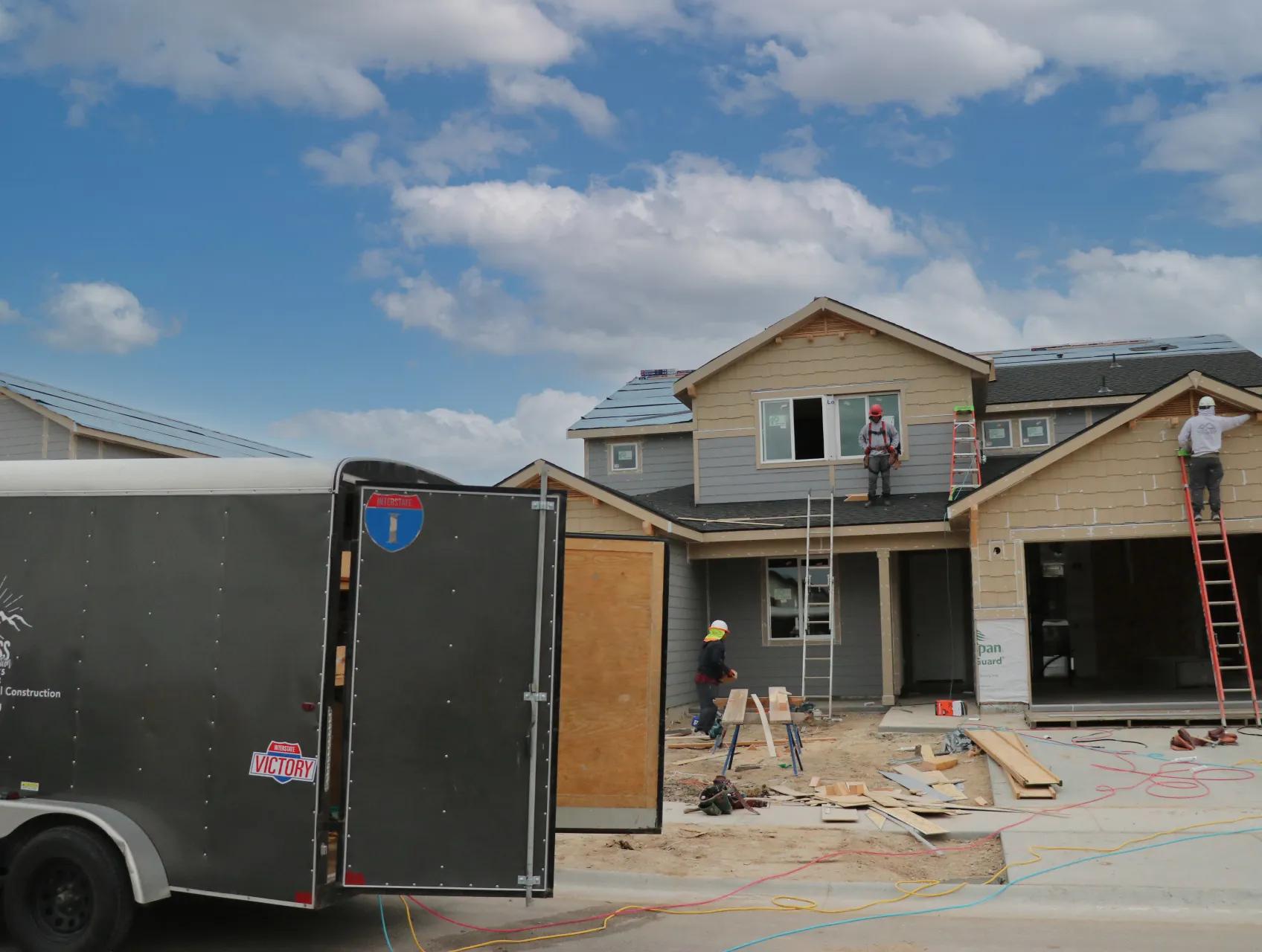 Workers in hard hats framing a two-story home with a small charcoal Interstate Victory cargo trailer parked at the jobsite curb