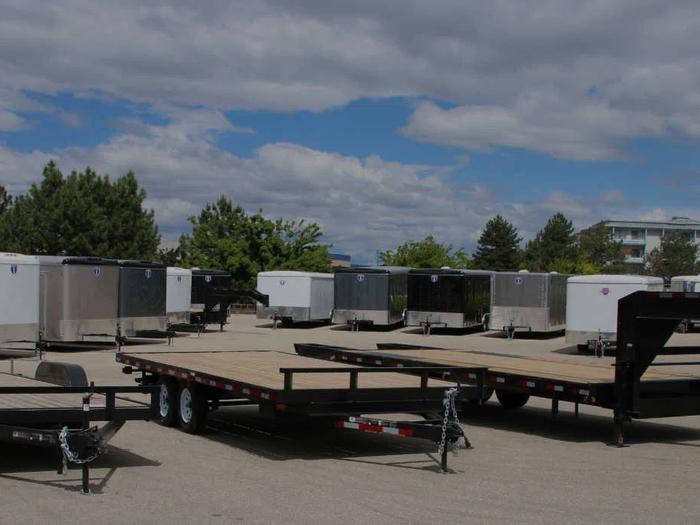 TrailersPlus dealer lot with two flatbed deckover gooseneck trailers in front of long rows of black and white enclosed cargo trailers