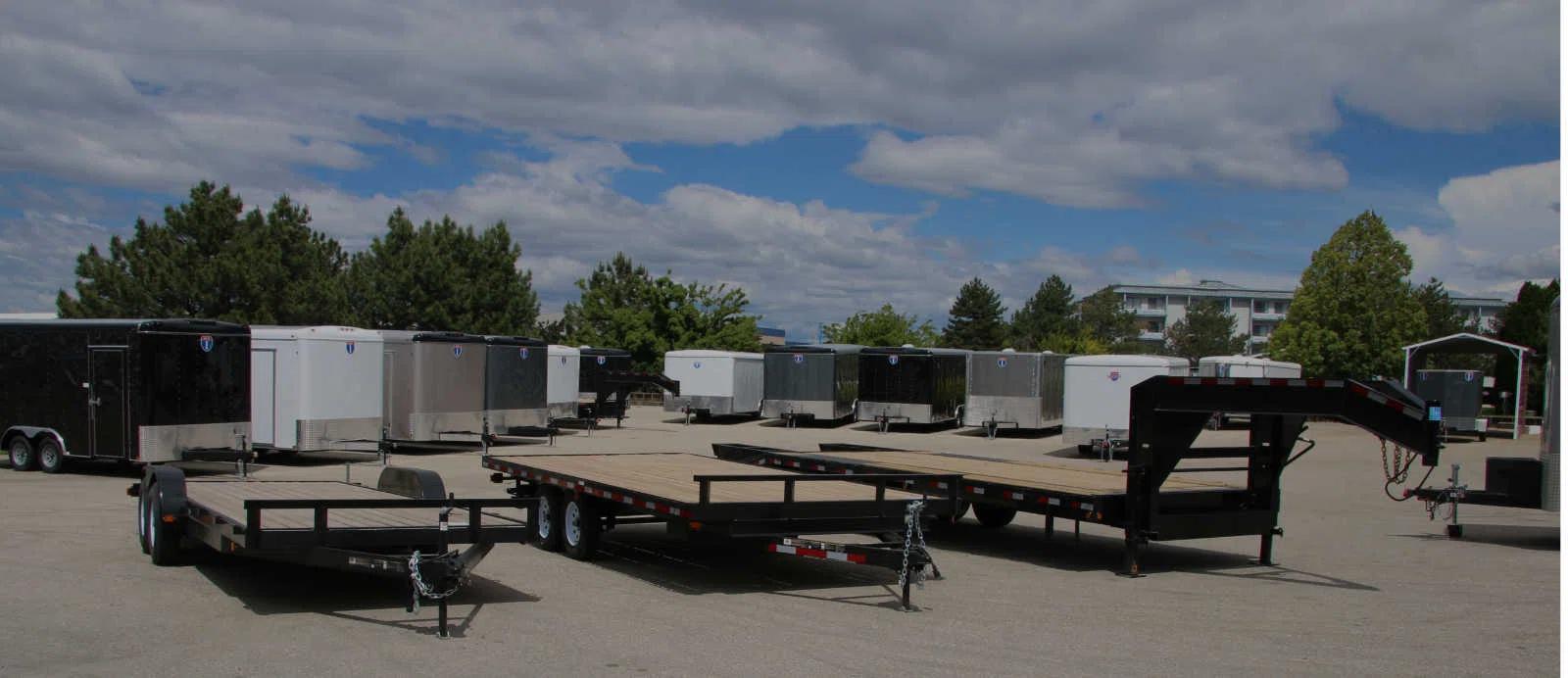 TrailersPlus dealer lot with two flatbed deckover gooseneck trailers in front of long rows of black and white enclosed cargo trailers