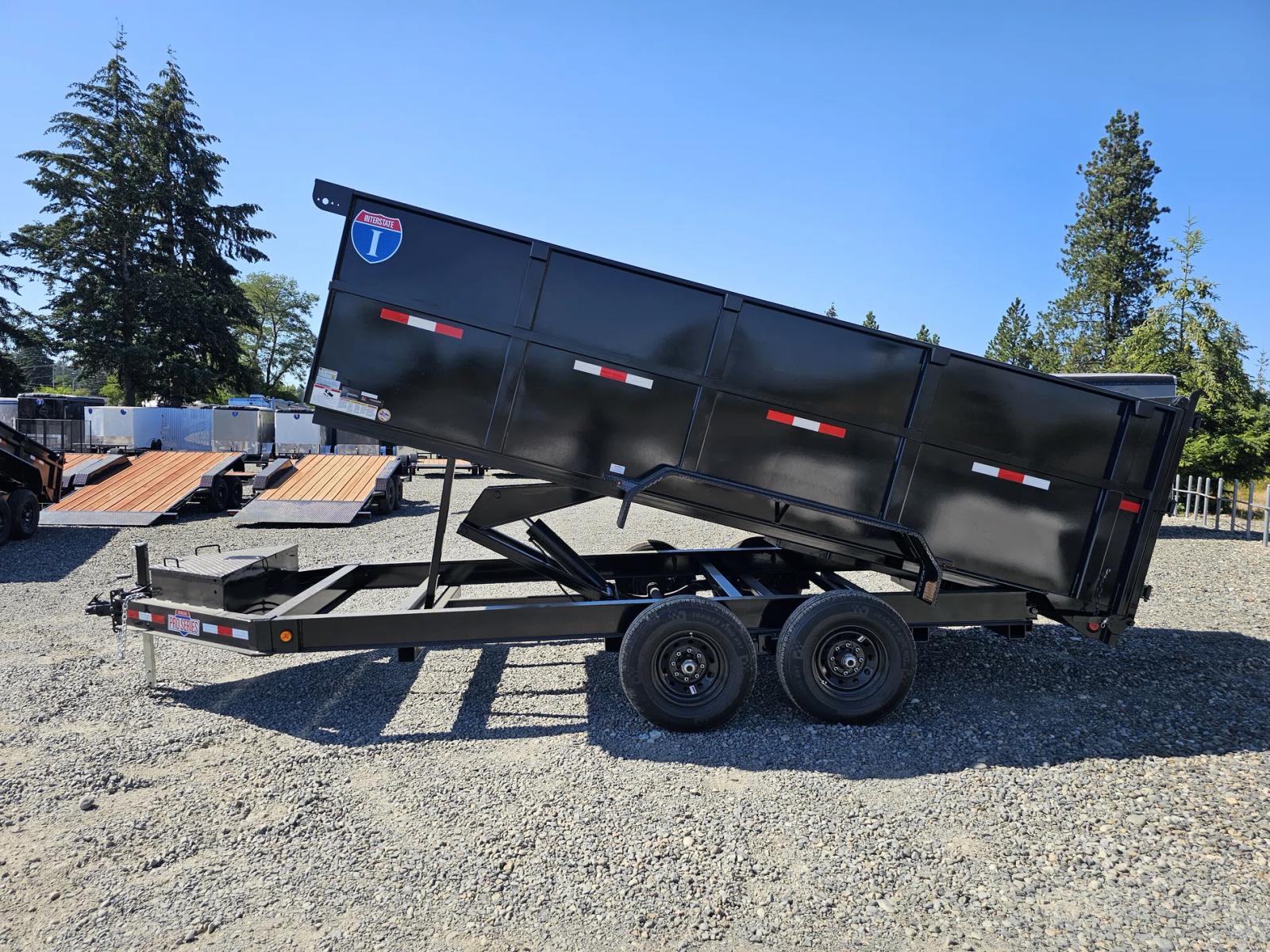 Black tandem-axle dump trailer with tall side extensions and the bed tilted up in dump position, parked at a gravel dealer lot