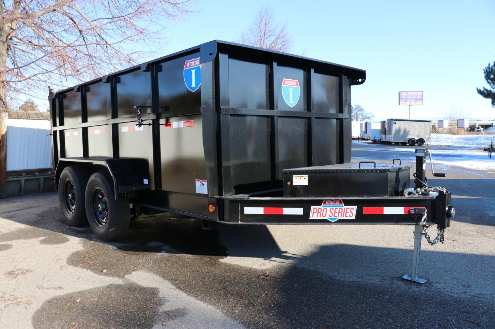 Black tandem-axle dump trailer with tall walls and a 'Pro Series' badge parked on asphalt next to a snowy edge of a dealer lot
