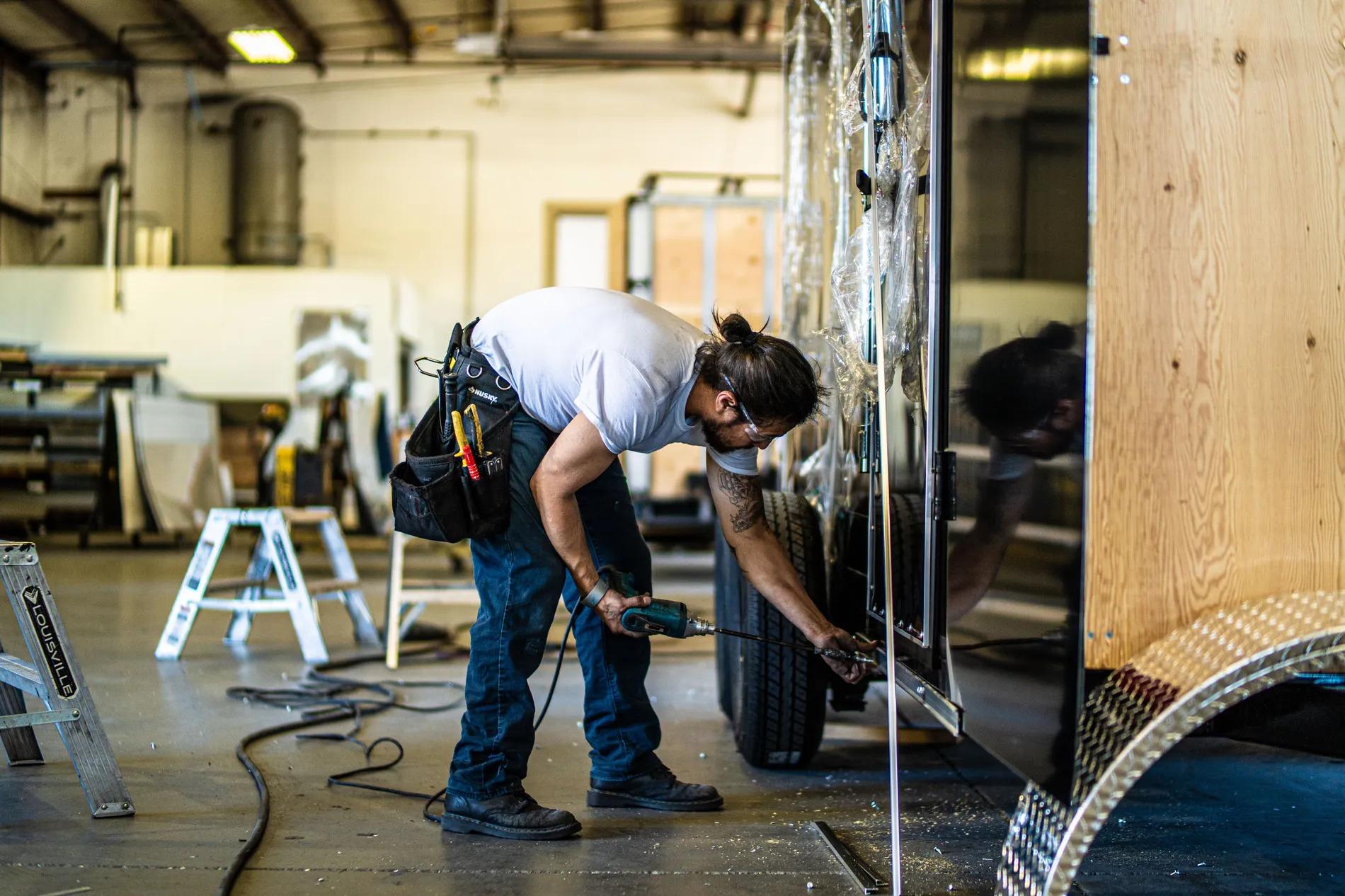 Bearded technician inside a TrailersPlus workshop using a power drill to service the side panel of an enclosed cargo trailer