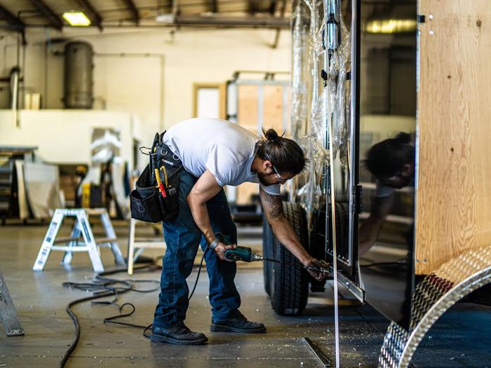 Bearded technician inside a TrailersPlus workshop using a power drill to service the side panel of an enclosed cargo trailer
