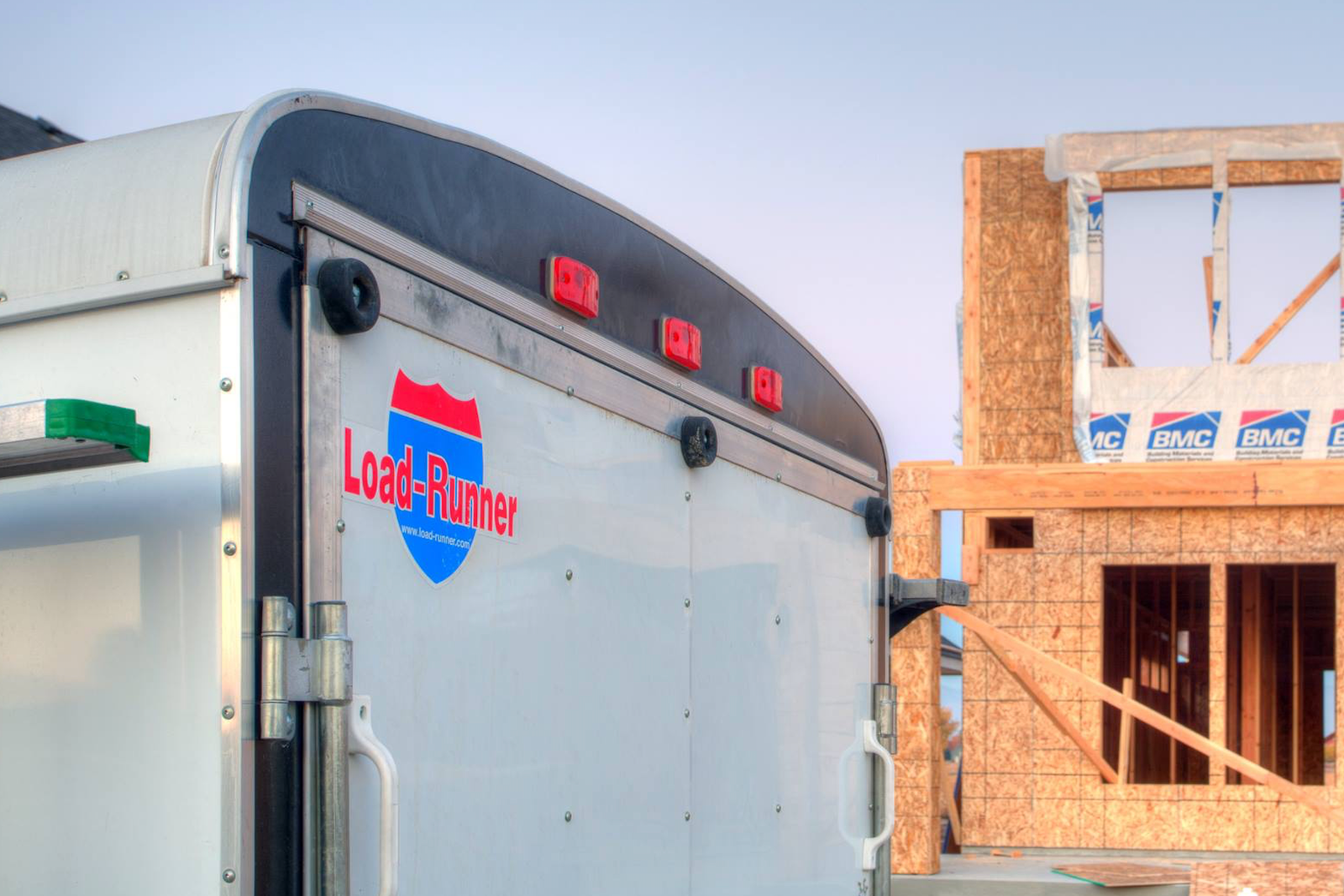 Rear of a white enclosed cargo trailer with a Load-Runner shield decal, parked beside a wood-framed house under construction