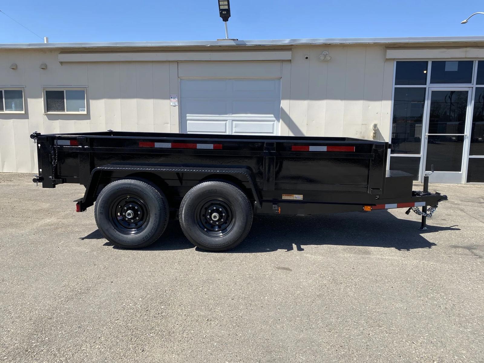 Side profile of a black tandem-axle dump trailer with closed sides parked beside a tan industrial building under a clear blue sky