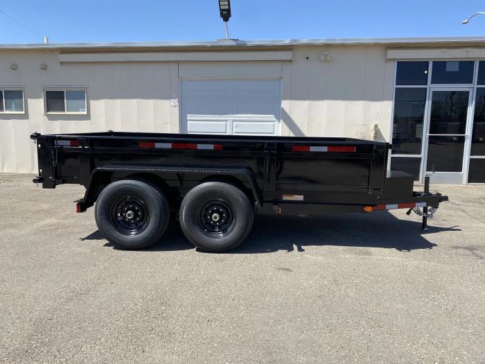 Side profile of a black tandem-axle dump trailer with closed sides parked beside a tan industrial building under a clear blue sky