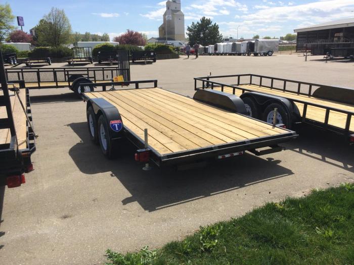 Black tandem-axle flatbed equipment trailer with wood deck parked at a TrailersPlus lot with a grain elevator in the distance